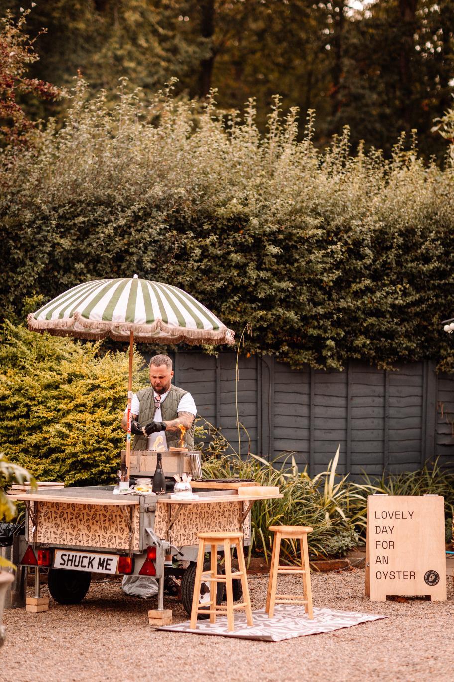 The O Co. trailer set up at a garden wedding with a green and white striped umbrella and a 'Lovely day for an oyster' sign